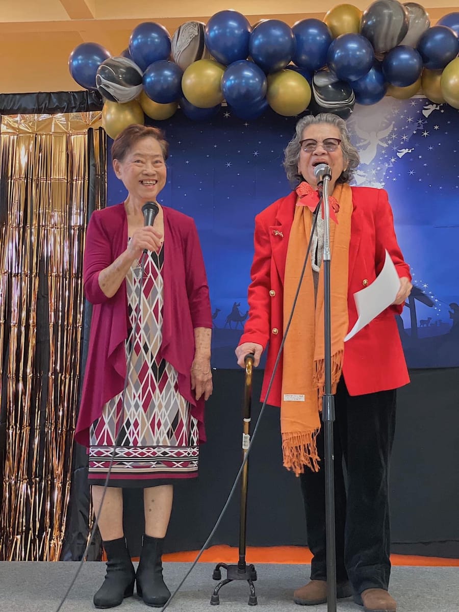 Two women singing into microphones under blue and gold balloons at a Christmas celebration
