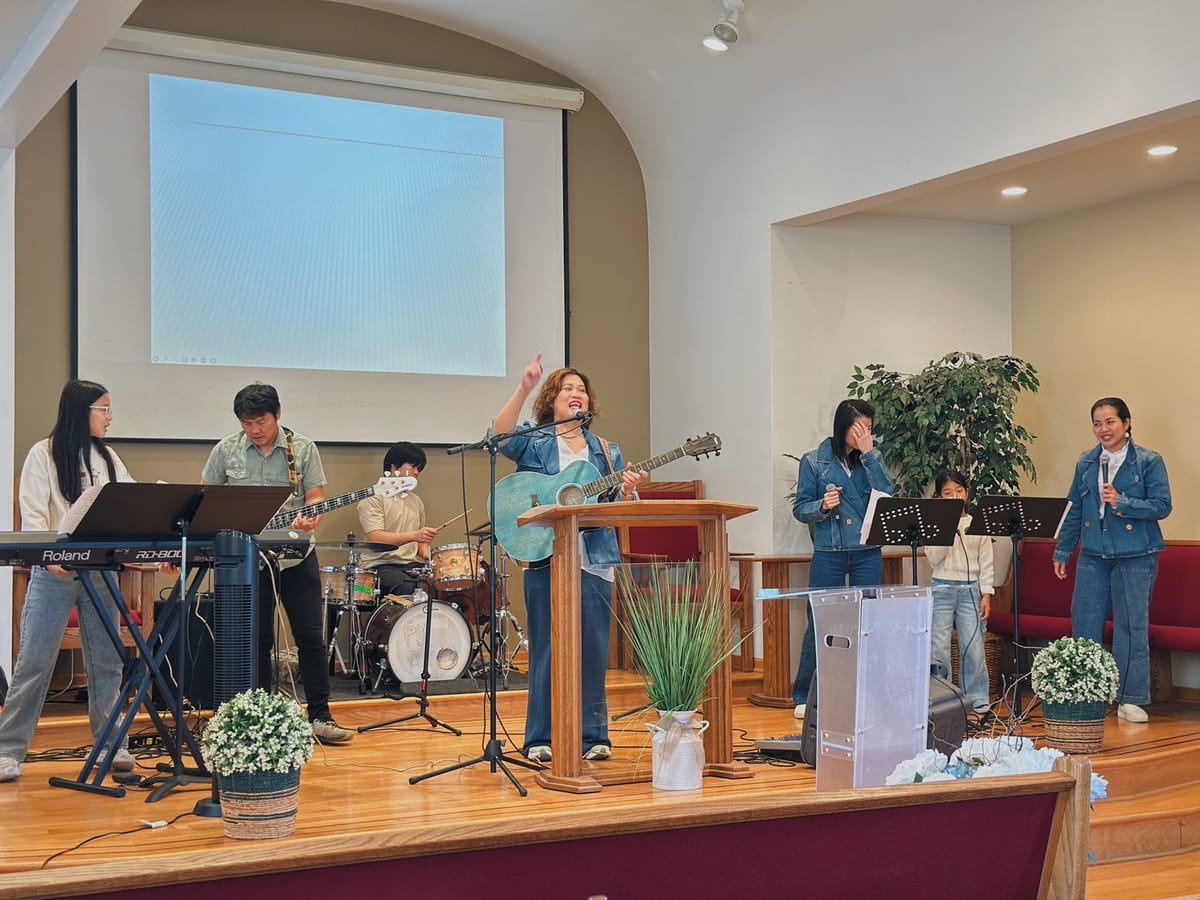 The worship band leading from the stage: a woman singing with an acoustic guitar at the lectern, keyboard, bass and drums behind her, and two singers with microphones to the right