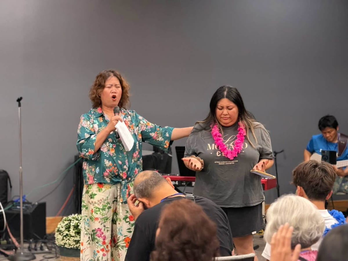 A woman holding a microphone prays with her hand on another woman’s shoulder while the congregation bows their heads